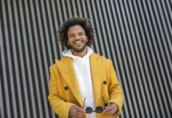 A cheerful Afro man in a yellow coat holds sunglasses, standing in front of a modern striped