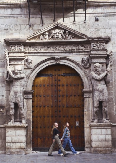 Two young woman friends casually walk past an ornate wooden door framed by intricate stone carvings
