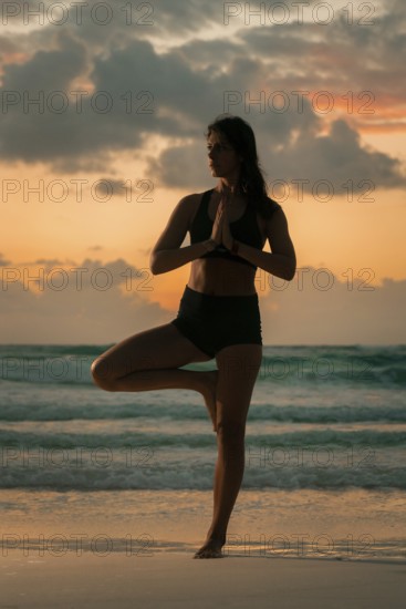A woman performs a yoga pose on a tranquil beach as the sun sets, blending calm and focus against a