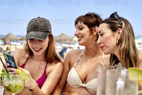 Three friends relax at a sunny beach, sipping colorful cocktails and checking their phones. The