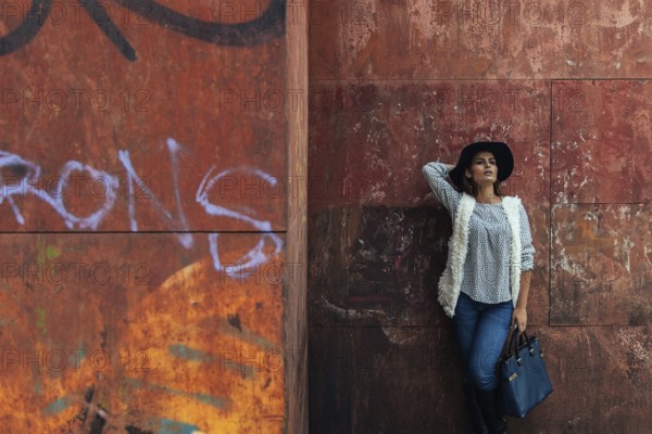 A stylish woman in casual attire with a hat leans against a graffiti-covered wall, showcasing urban