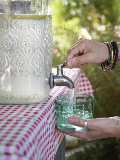 Fresh lemonade in large glass bottle with tap. Hands serving in glass. Refreshing summer drink.