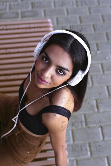 A young woman wearing white headphones sits on a wooden bench, looking up with a serene expression.