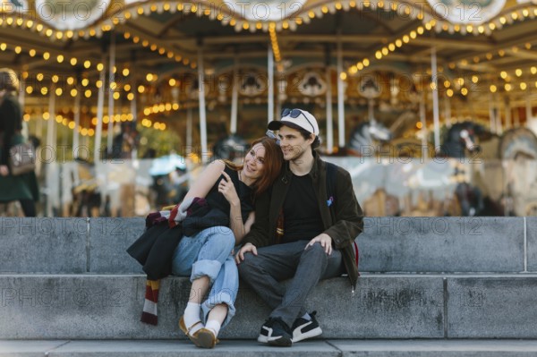 A couple sits on steps, embracing in front of a lit up carousel in Brooklyn Bridge Park. The joyful