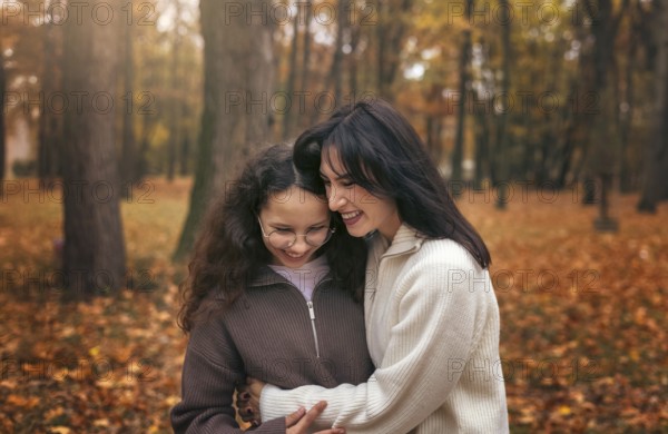 A mother and daughter share a warm embrace surrounded by autumn leaves in a forest. The cozy moment