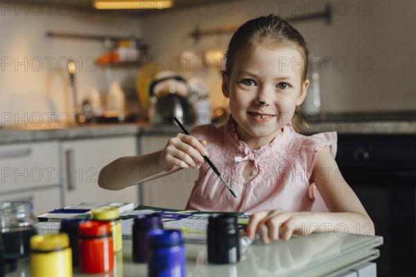 A joyful child with a painted nose engages in a creative painting activity at home. She holds a