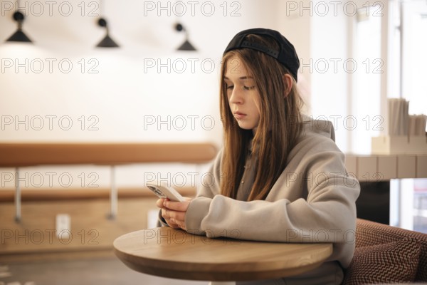 Teenage girl sitting at a cafe table using her smartphone. She is wearing a casual hoodie and cap,
