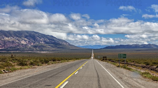 Endless road runs straight ahead through a dry landscape with mountains in the background and