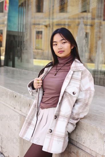 Young woman with a stylish plaid coat and pink blouse leaning against a stone ledge in an urban