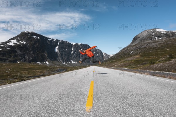 A traveler in a red jacket leaps mid-air on an open mountain road in Norway Framed by dramatic