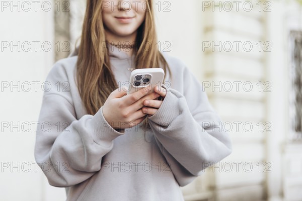 Teenage girl holding a smartphone while outdoors, dressed in a casual gray sweatshirt. The focus is