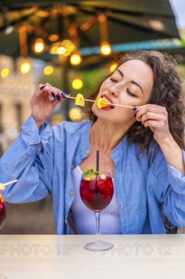 Vertical photo of a caucasian woman enjoying biting the citrus garnishing a cocktail in an outdoor