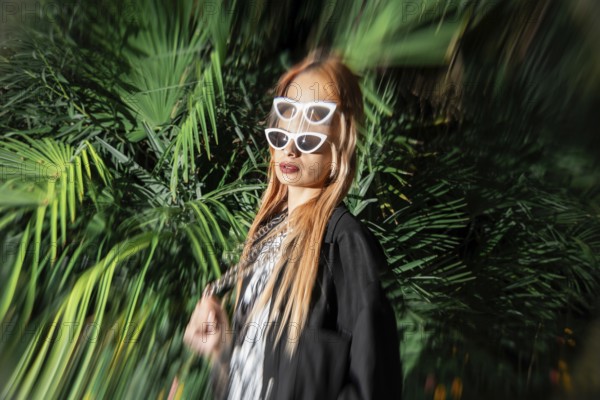 A stylish woman with double sunglasses and long hair stands confidently in front of vibrant green