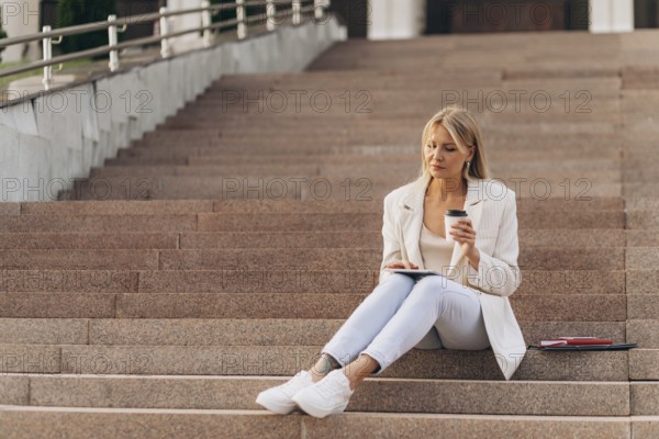 A businesswoman in casual business attire sits on outdoor steps, holding a coffee cup and using a