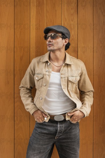A fashionable man wearing sunglasses, a tan jacket, and a cap stands confidently against a wooden