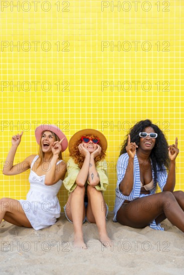 Three diverse women wearing summer clothes and hats are sitting on the sand, pointing upwards with