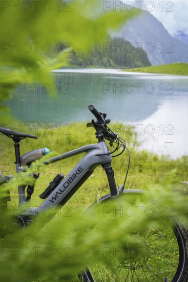 An e-bike is parked on the lakeshore. The surrounding nature is lush and green, and the mountains