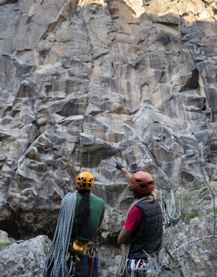 Two climbers equipped with helmets and ropes assess a towering rock face for a traditional climbing