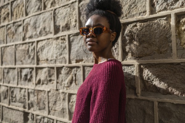 A stylish woman in a burgundy sweater wearing sunglasses stands confidently against a textured