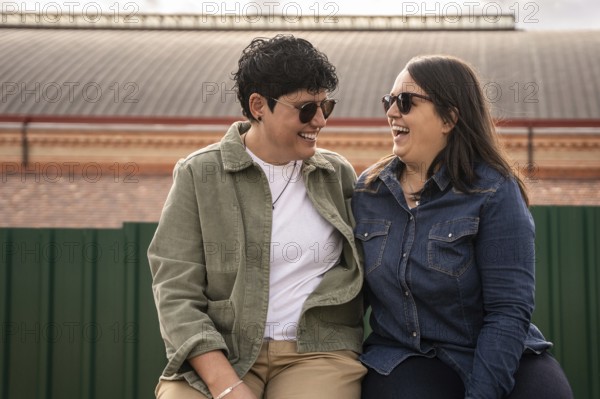 A joyful curvy lesbian couple shares a light heart moment outdoors, sitting closely and wearing