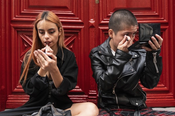 A lesbian couple sits together in front of a red door, each applying makeup with focus and care,