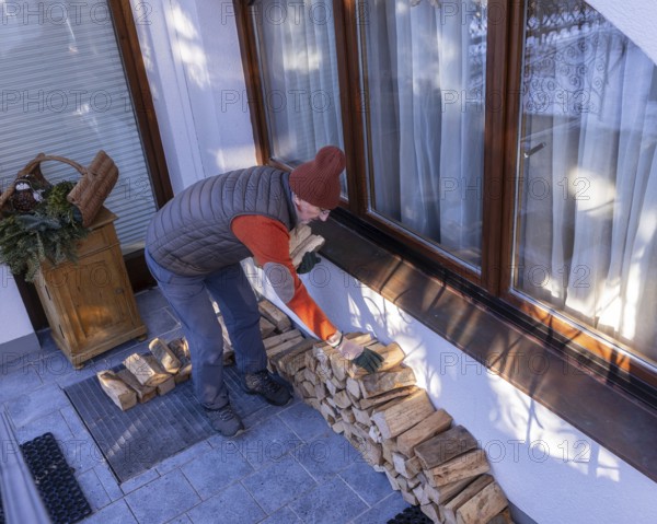 A senior man in casual winter wear stacks eco-friendly firewood next to his home on a clear day.