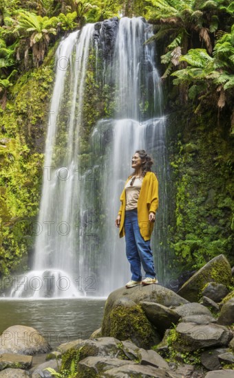 A woman stands calmly on a rock, surrounded by the lush greenery of a forest with a stunning