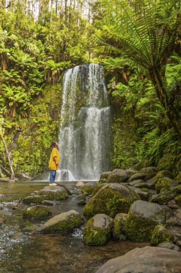 A woman stands by a waterfall in a lush forest, surrounded by moss covered rocks and vibrant ferns,