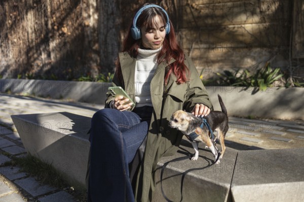 A young woman sits on a bench with headphones, holding a smartphone, while petting her small