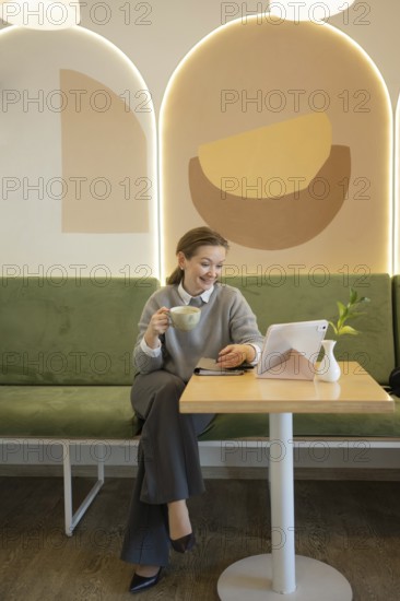 A woman relaxes in a stylish cafe, sipping coffee and using a tablet. The tranquil setting is