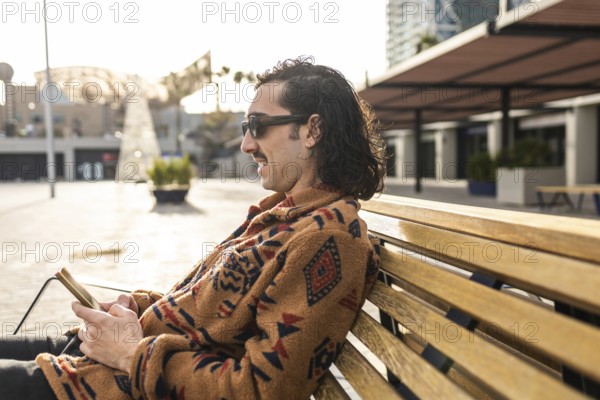 A man sits calmly on a bench outdoors with a smartphone Wearing sunglasses and a stylish patterned