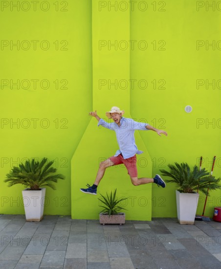 Young man jumping over plant pot in front of green house wall, green house facade with flowers,