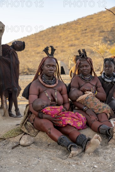 Married Himba woman with their babies in their arms sitting in front of the hut of the first woman,