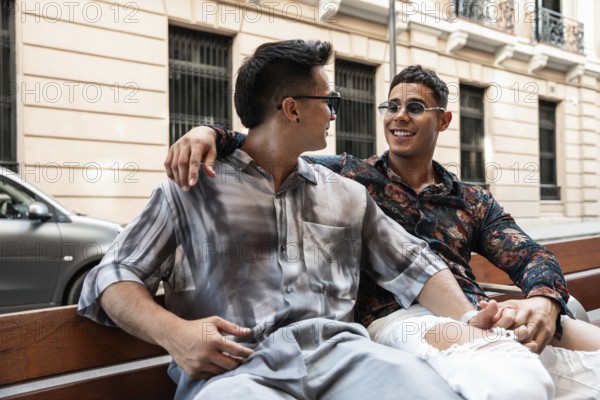 A cheerful latin gay couple sits close together on a bench, laughing and enjoying a sunny day in