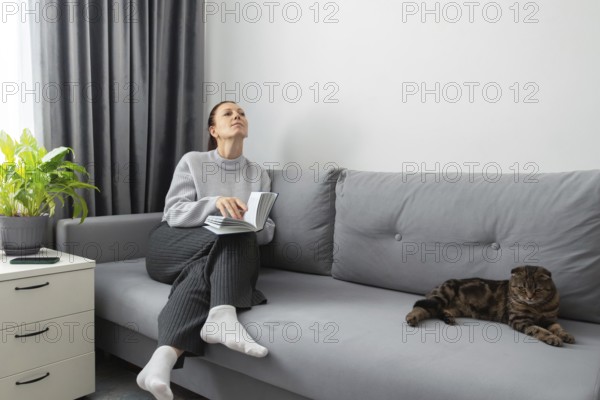 A woman sits on a gray sofa, holding a book, looking peaceful and contemplative. Beside her, a cat