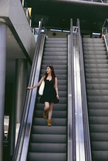 A young asian woman in a black dress and boots descends an escalator in an urban setting. She