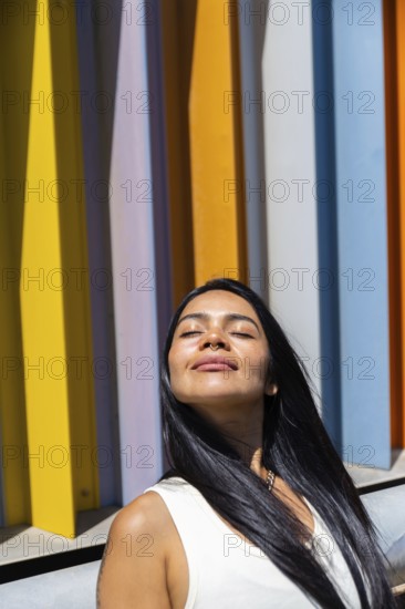 A woman in a colorful outfit enjoys the sunlight, standing in front of a backdrop of vibrant,