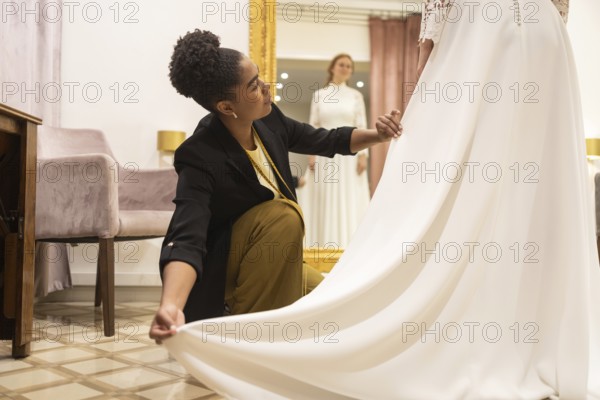 A seamstress carefully adjusts a flowing white bridal gown for a customer in a stylish bridal