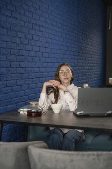 A woman with glasses sits at a cafe table with a laptop, tea cup, and teapot, against a stylish