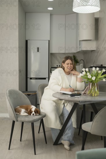 A woman sits at a modern kitchen table, writing in a notebook. Nearby, a small dog rests on a chair