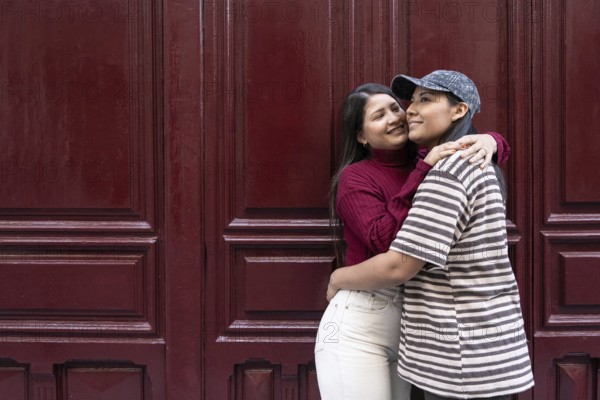A Latin lesbian couple shares a loving embrace against a deep red wooden backdrop. They exude