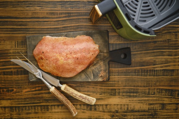 A piece of roasted pork meat on a wooden cutting board. Kitchen knife and fork are placed next to