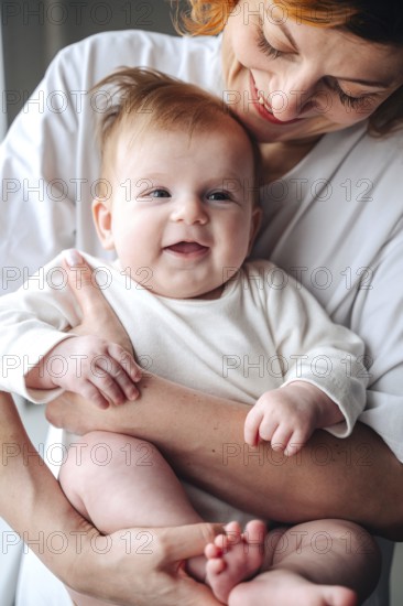 Loving mother cradles smiling newborn baby girl at home, both wearing white clothes. The baby's