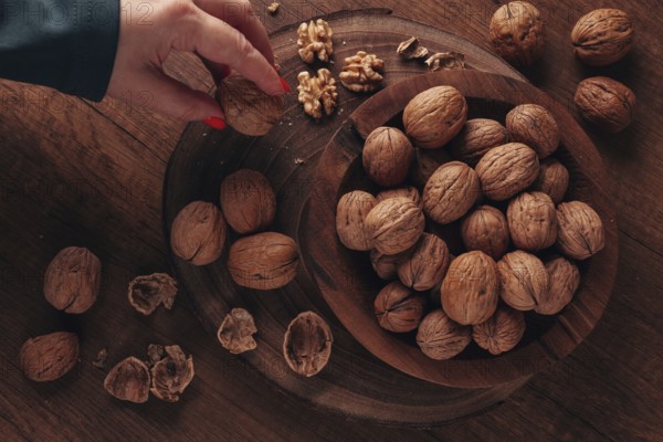 A woman's hand taking a walnut from a wooden bowl filled with walnuts. There are shells and walnuts