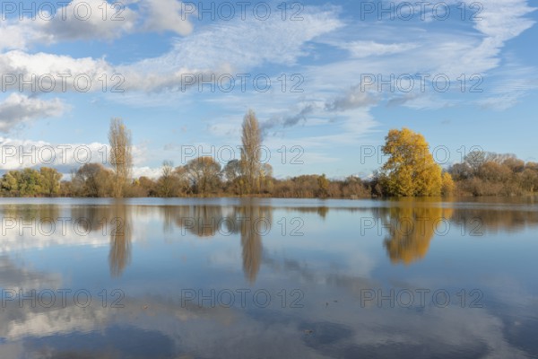 The river flows flat and reflects clouds and mountains under a clear sky. Autumn trees draw the
