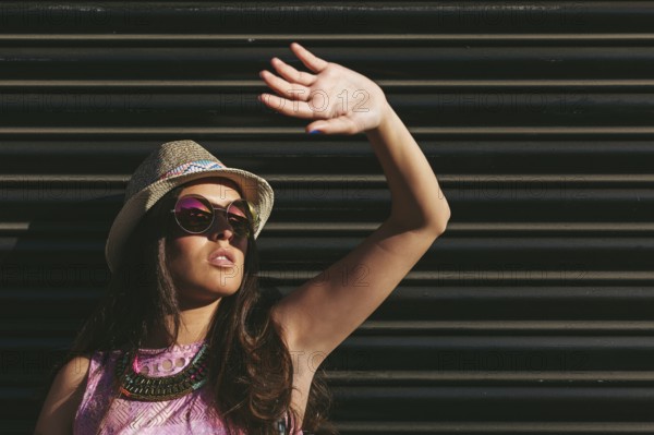 Woman wearing a summer hat and sunglasses raises hand to shield eyes from the sun. The background