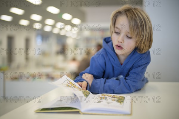 Interior shot, boy, 6 years old, blonde, reading book in the children's section of the city
