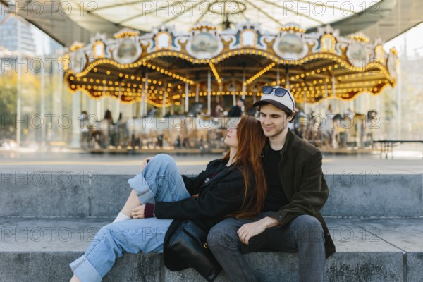 A young couple sits on steps in front of a brightly lit carousel in Brooklyn Bridge Park. They
