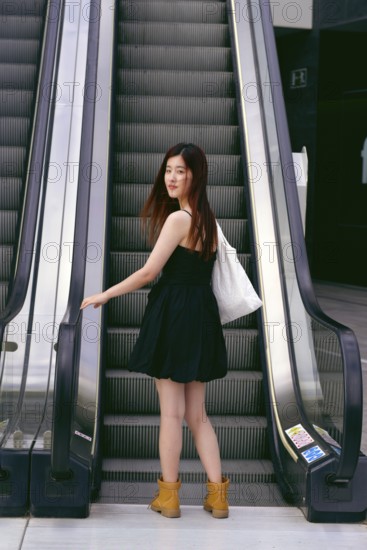 A young asian woman in a black dress and boots stands on an escalator in an urban setting, holding