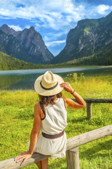 Woman wearing straw hat admiring lake dobbiaco with the dolomites in the background on a sunny
